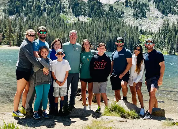 A large group of people of varying ages smiling at the camera, standing by a clear blue lake with a dense pine forest and … A large group of people of varying ages smiling at the camera, standing by a clear blue lake with a dense pine forest and ...